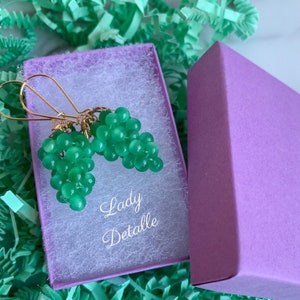 May include: A pair of green grape earrings in a purple box. The earrings are made of small green beads and have gold hooks. The box has the text "Lady Detalle" on it.