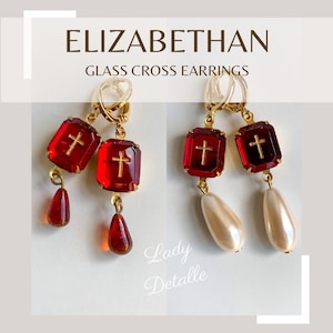 May include: Gold cross earrings with red glass cross charms and dangling teardrop-shaped beads. The earrings are labelled "ELIZABETHAN GLASS CROSS EARRINGS" and "Lady Detalle".