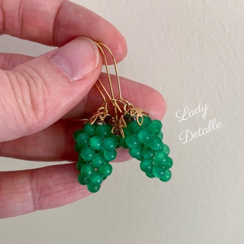 May include: A pair of green glass bead earrings shaped like grapes with gold-toned metal accents. The earrings are hanging from gold-toned hooks.