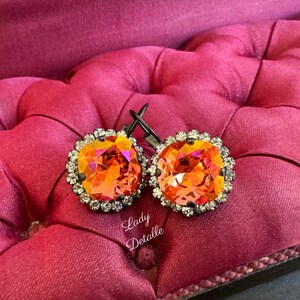May include: A pair of orange crystal earrings with a silver-colored frame and a halo of small clear crystals. The earrings are displayed on a pink, tufted surface. The text "Lady Detalle" is visible.