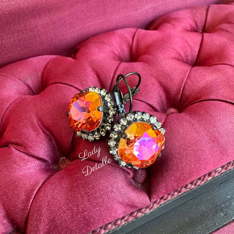 May include: A pair of orange and crystal earrings with a black metal setting. The earrings have a large, faceted orange center stone surrounded by smaller crystals. The text "Lady Detalle" is visible in the image.