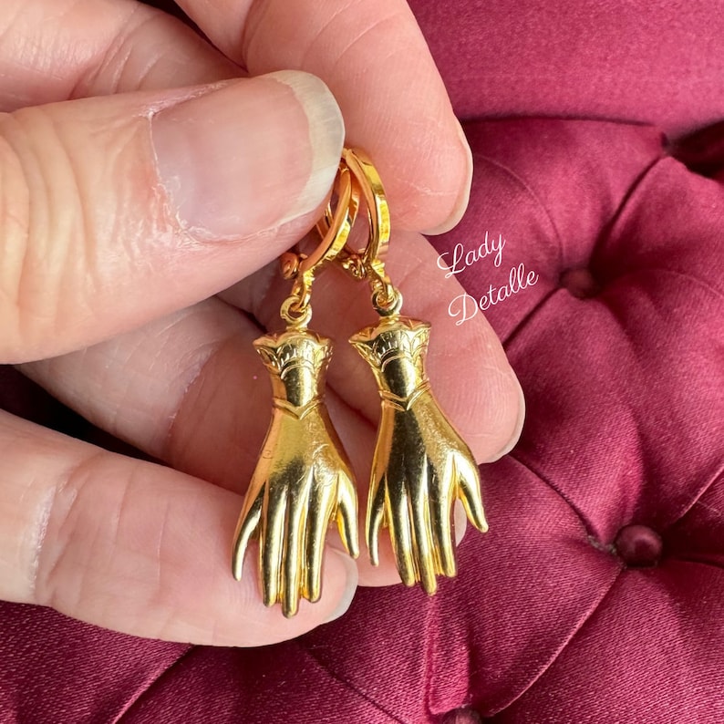 K&ouml;nnte beinhalten: Goldene Creolen mit einem baumelnden Hand-Anh&auml;nger. Die H&auml;nde sind detailliert und haben eine strukturierte Oberfl&auml;che.