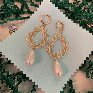 May include: A pair of gold earrings with a delicate leaf design and a pearl drop. The earrings are on a white background.