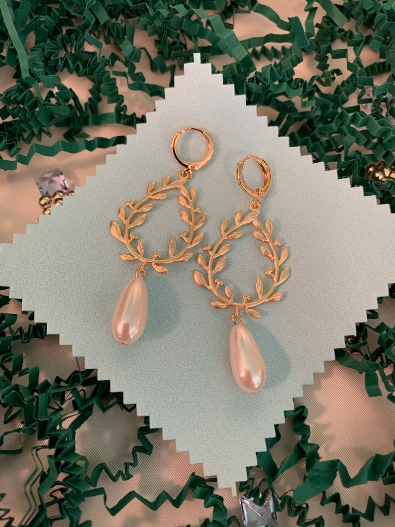 May include: A pair of gold earrings with a delicate leaf design and a pearl drop. The earrings are on a white background.