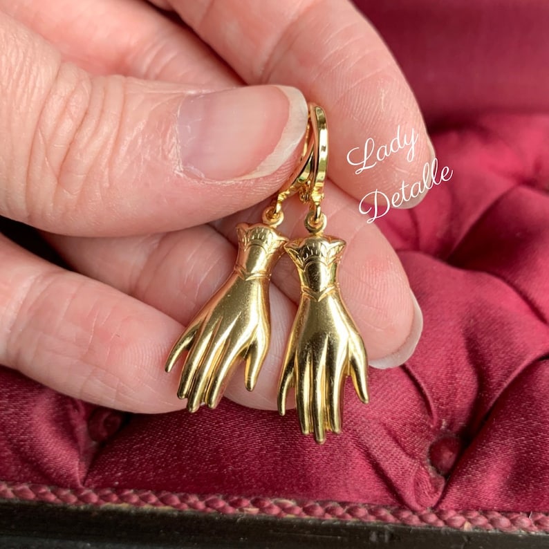 K&ouml;nnte beinhalten: Goldfarbene Ohrringe mit einem Paar H&auml;nden mit einem detaillierten Design. Die Ohrringe h&auml;ngen an einem Ring.