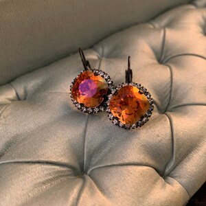 May include: A pair of orange and iridescent crystal earrings with a square shape. The earrings are surrounded by a ring of small, clear crystals and have dark metal lever-back closures. The earrings are displayed on a light green, tufted surface.
