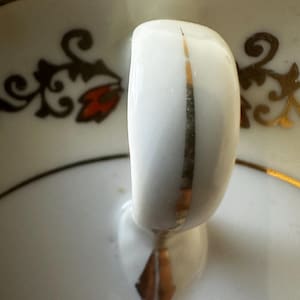 Puede incluir: Una taza de té blanca con un borde dorado y un diseño floral. La taza tiene un asa con una franja dorada.