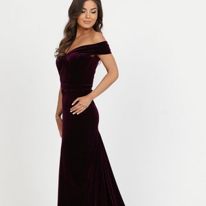 May include: A long, burgundy velvet dress with an off-the-shoulder neckline and a fitted bodice. The dress has a flowing train.
