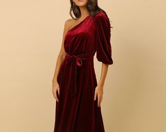 May include: A long, one-shoulder burgundy velvet dress with a puff sleeve and a matching sash tied at the waist.