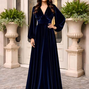 A full-length navy blue velvet dress with a V-neck and long balloon sleeves. The dress features a fitted bodice and a flowing a line maxi skirt.