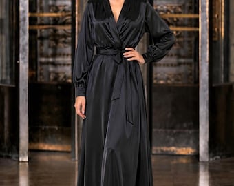 May include: A floor-length black satin wrap dress featuring long sleeves with cuffed wrists, a deep V-neckline, and a matching fabric tie belt at the waist. The garment has a smooth, reflective finish and a flowing skirt that reaches the floor.
