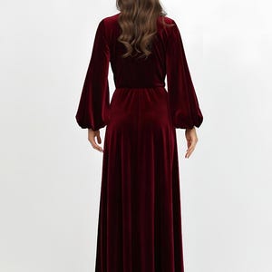 A long, burgundy velvet dress with a V-neck and long, voluminous sleeves. The dress features a fitted waist and a flowing skirt. The model is seen from the back, highlighting the dress's elegant design and rich color. The dress is made of a soft velvet material.