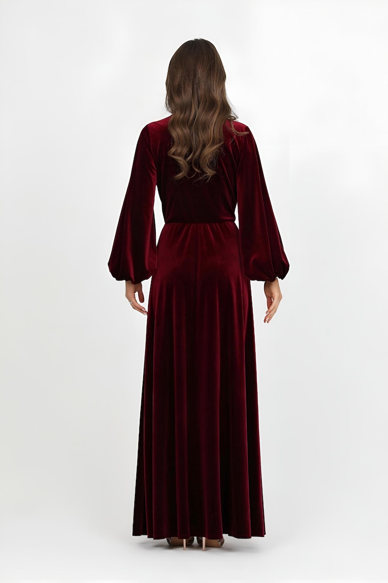 A long, burgundy velvet dress with a V-neck and long, voluminous sleeves. The dress features a fitted waist and a flowing skirt. The model is seen from the back, highlighting the dress's elegant design and rich color. The dress is made of a soft velvet material.