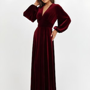 A full-length, burgundy velvet dress featuring a V-neck and long sleeves. The dress has a fitted waist and a flowing skirt. The sleeves are gathered at the wrist. The dress is made of a soft, luxurious fabric.