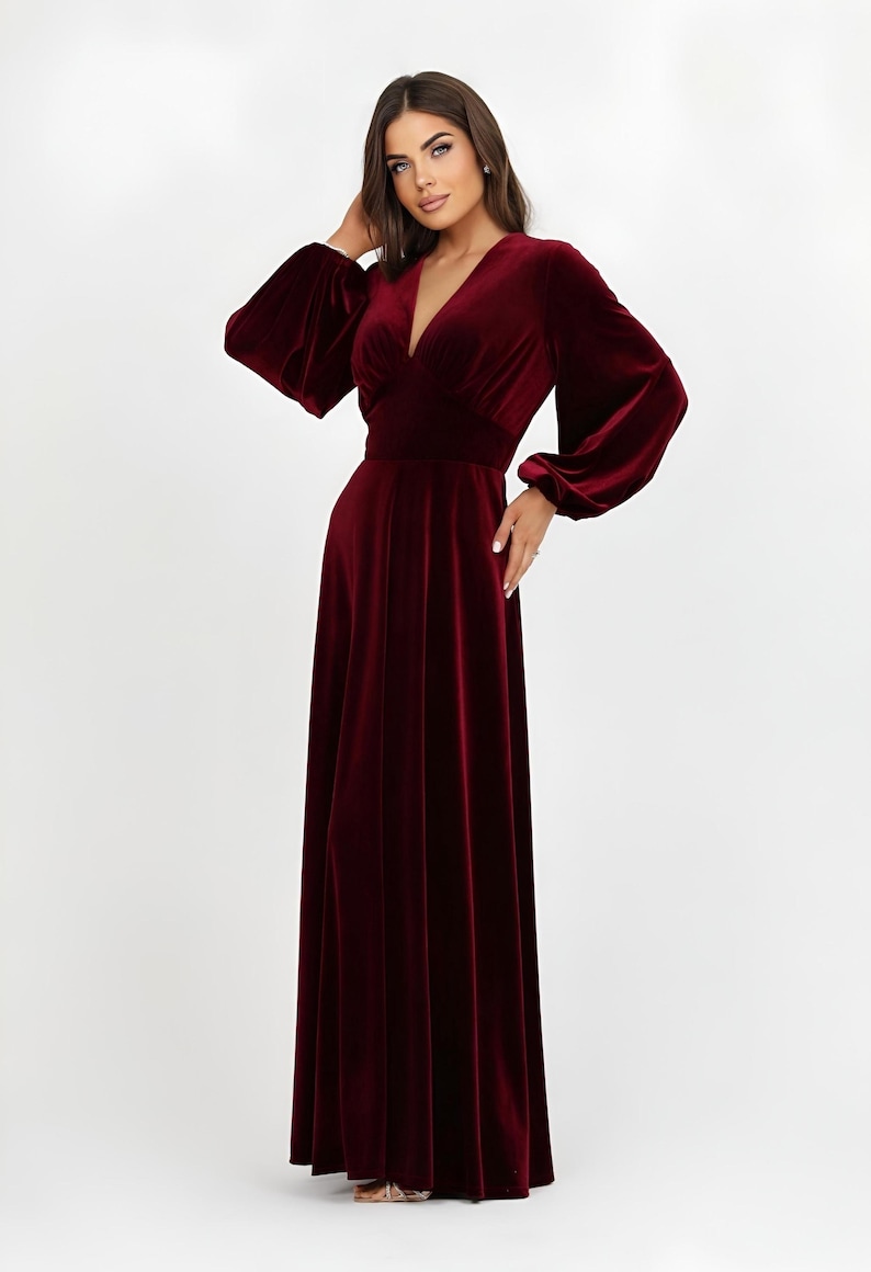 A full-length, burgundy velvet dress featuring a V-neck and long sleeves. The dress has a fitted waist and a flowing skirt. The sleeves are gathered at the wrist. The dress is made of a soft, luxurious fabric.