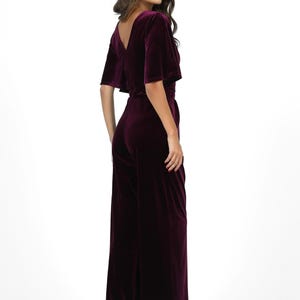 May include: Plum-colored velvet jumpsuit with a V-neckline and wide-leg pants. The short-sleeved top has a slight flutter detail. The jumpsuit is cinched at the waist and worn with black high-heeled shoes. The model is facing away from the camera.