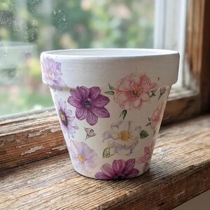 May include: A white ceramic plant pot adorned with watercolour floral patterns in shades of purple and pink. The pot has a cylindrical shape with a slightly flared rim and is on a wooden surface near a window.