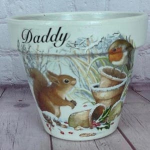 Könnte beinhalten: Cremefarbener Keramik-Blumentopf mit einer Winterszene mit einem Eichhörnchen und einem Rotkehlchen. Der Topf trägt den Schriftzug "Daddy" in schwarzer Schrift. Das Design umfasst Schnee, Pflanzen und einen kleinen umgestürzten Topf.