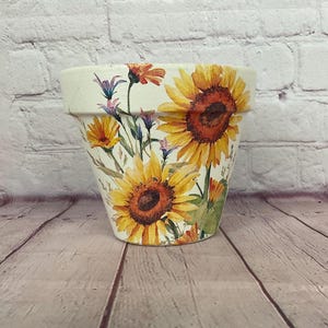 May include: A white ceramic flower pot decorated with large yellow sunflowers and smaller orange flowers. The pot has a wide rim and is set against a light-colored brick wall and wooden surface.