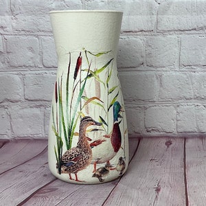 May include: White ceramic vase with a floral design featuring ducks and cattails. The vase has a wide base and a narrow neck.