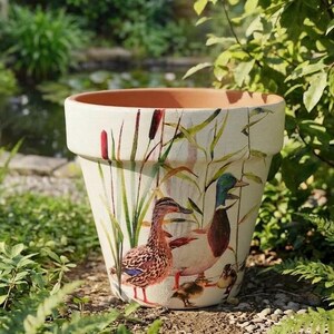 May include: A large, off-white ceramic plant pot with a painted duck scene. The pot features a family of ducks amidst reeds and cattails. The rim is terracotta, and the pot is set outdoors.