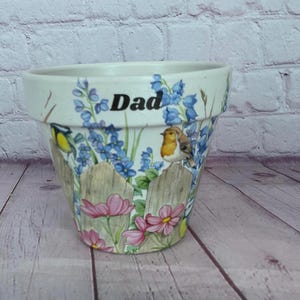 Könnte beinhalten: Weißer Keramik-Blumentopf mit einem bemalten Blumendesign. Der Topf zeigt blaue Blumen, rosa Blumen und Vögel. Das Wort "Dad" steht oben. Der Topf ist mit einem Holzzaun verziert.