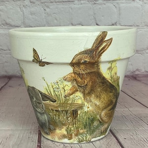 Pode incluir: Um vaso de flores de cerâmica branca com um desenho pintado com um coelho marrom, um coelho cinza e uma borboleta. O vaso tem um aspecto ligeiramente envelhecido, com uma estética rústica.