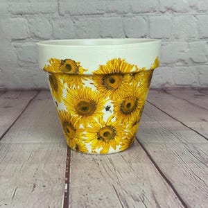 Puede incluir: Una maceta de cerámica blanca y amarilla decorada con un patrón de girasoles. La maceta presenta un borde ancho y está cubierta de girasoles amarillos brillantes con algunas abejas. La maceta está sobre una superficie de madera.