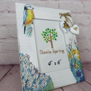 Puede incluir: Marco de fotos de madera blanca con temática primaveral. El marco tiene un pájaro azul, flores y mariposas. El marco mide 10,16 cm por 15,24 cm y dice "Nicola Spring" en la parte delantera.