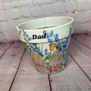 Può includere: Vaso di fiori in ceramica bianca con un design floreale con campanule, fiori rosa e uccelli. Il vaso è personalizzato con la parola "Papà" in lettere dorate.