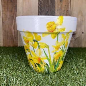 May include: White ceramic flower pot decorated with yellow daffodils. The pot has a wide rim and a tapered body. The daffodils are painted with yellow petals and green stems and leaves. The pot is sitting on green artificial grass.