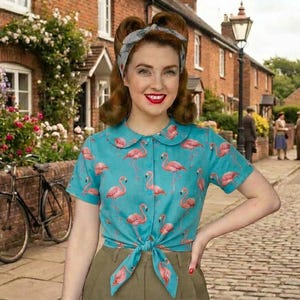 Peut inclure: Une femme porte un chemisier turquoise à manches courtes orné de flamants roses, noué à la taille. Elle se tient sur une rue pavée de briques devant des bâtiments et un vélo.