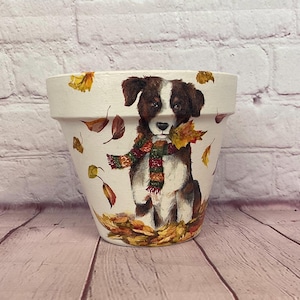 Puede incluir: Maceta de cerámica blanca con una ilustración pintada a mano de un perro con una bufanda y una hoja. La maceta está decorada con hojas de otoño caídas en tonos amarillos, naranjas y marrones.
