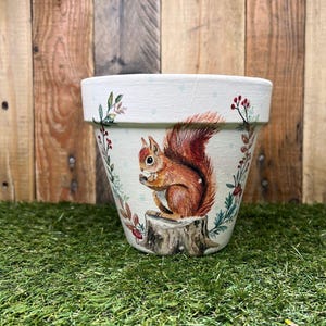 Puede incluir: Maceta de cerámica color crema con una ardilla roja pintada a mano sobre un tocón. La maceta está decorada con detalles florales y de bayas. Adecuada para uso en interiores o exteriores, ideal para plantas.