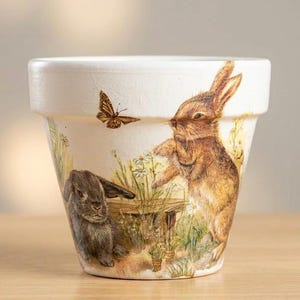 Puede incluir: Maceta de cerámica blanca con una escena pintada de dos conejos en un jardín. Un conejo está de pie, extendiendo la mano hacia una mariposa. El otro conejo está sentado cerca de una pequeña mesa de madera. La maceta tiene un borde ancho y una forma ligeramente cónica.