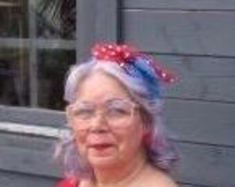 May include: A woman with white and purple hair wearing a red shirt and a red, white, and blue polka dot bow in her hair.