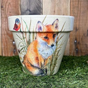 May include: A white ceramic planter featuring a painted image of a fox in a grassy field. The fox is orange and white with a black nose and black eyes. A small bird is perched on a branch in the background.