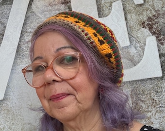 May include: A colorful, knitted beret in shades of brown, yellow, green, and red. The hat is worn by a person with purple hair and glasses. The person is wearing a black and white polka dot top. The background includes white letters and a gold garland.