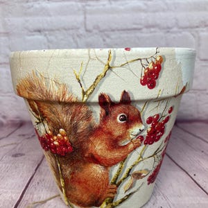 Puede incluir: Maceta de cerámica decorativa con una ilustración detallada de una ardilla comiendo bayas rojas. La maceta tiene un fondo crema con ramas y bayas. La ardilla es marrón y naranja, añadiendo un toque de naturaleza a la decoración del hogar.