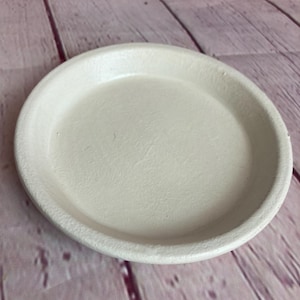 Peut inclure: Une assiette ronde en céramique blanc cassé avec un bord légèrement surélevé. L'assiette a une surface lisse et convient pour servir des aliments ou comme objet de décoration. L'assiette est présentée sur un fond en bois rose et marron.