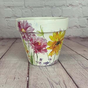 Puede incluir: Una maceta de cerámica blanca decorada con coloridos diseños florales. La maceta presenta flores grandes y vibrantes en tonos rosa, amarillo y morado, con tallos y hojas verdes. La maceta está sobre una superficie de madera.