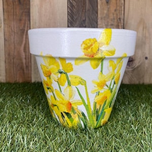 Puede incluir: Maceta pintada de blanco con un diseño pintado a mano de narcisos amarillos y tallos verdes. La maceta está sobre un lecho de césped artificial verde. La maceta mide aproximadamente 15 cm de alto.