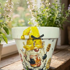 May include: A white ceramic plant pot with a gnome design wearing a yellow hat and holding a honey jar. The pot is decorated with bees and a honeycomb. Ideal for flowers or herbs, it measures approximately 15 cm in height.