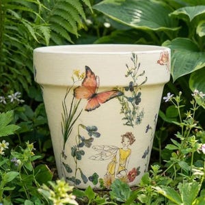 May include: Cream-colored ceramic flower pot decorated with a butterfly, fairy, and floral illustrations. The pot is surrounded by green foliage and flowers, creating a garden theme. The pot is a standard size for planting.