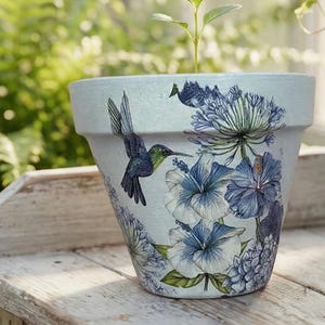 Könnte beinhalten: Hellblauer Keramik-Blumentopf mit einem Kolibri- und Blumenmuster. Der Topf zeigt blaue und weiße Hibiskusblüten und andere blaue florale Elemente. Eine kleine Pflanze wächst aus der Oberseite des Topfes.