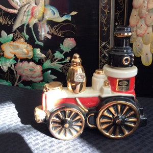 May include: A red, white, and black ceramic train engine decanter with gold accents. The engine has a black chimney and a gold-colored bottle on top. The decanter is sitting on a gray surface.