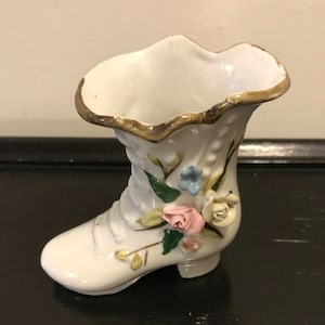 May include: A white porcelain boot with a ruffled top and floral accents. The boot is decorated with pink, white, and blue flowers and green leaves.