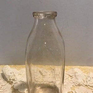 May include: A clear glass milk bottle with a rounded bottom and a slightly flared top. The bottle is empty and has a vintage look.