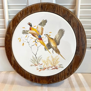 May include: A round wooden framed trivet with a white ceramic tile featuring a colorful pheasant and a brown pheasant in a woodland setting.