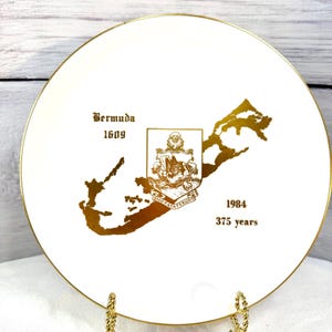 May include: A white decorative plate with a gold rim, featuring a gold map of Bermuda and text that reads "Bermuda 1609" and "1984 375 years". The plate is displayed on a gold stand.